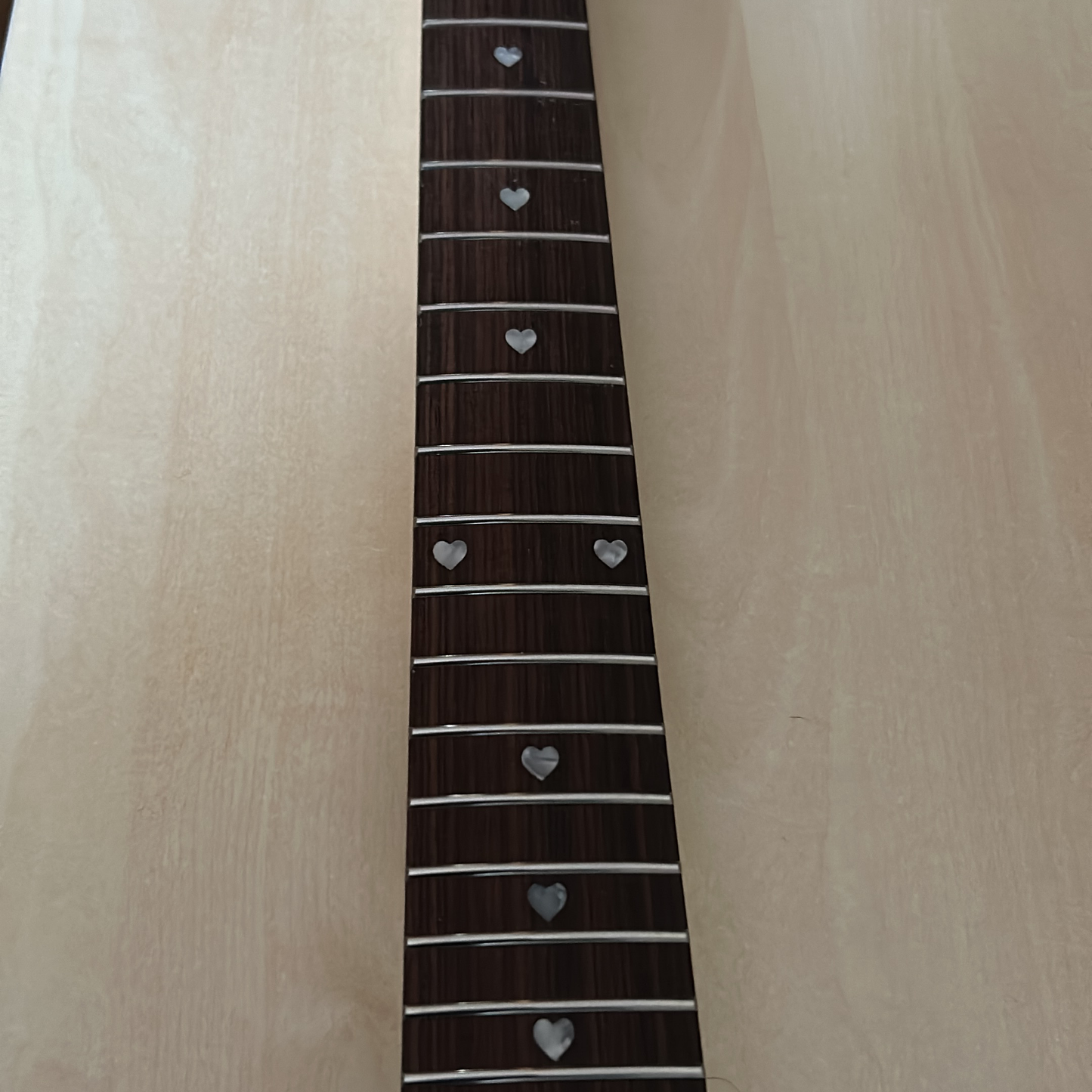 Close-up of a guitar fretboard with dark brown wood, silver metal frets, and silver heart-shaped inlays, shown against a light wood background.