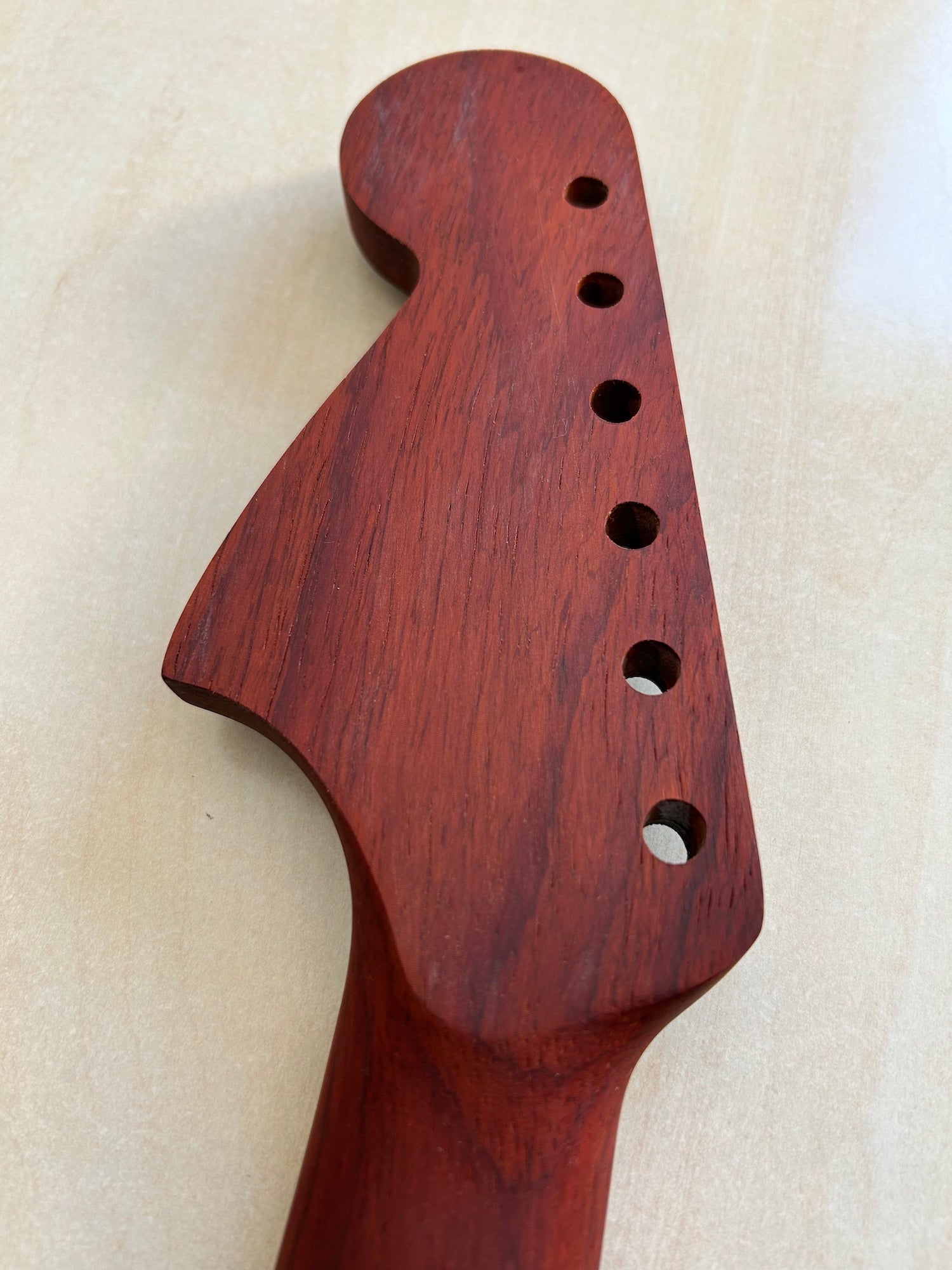 Close-up of a reddish-brown wooden guitar headstock neck joint with six tuners and visible wood grain, photographed on a light beige surface.