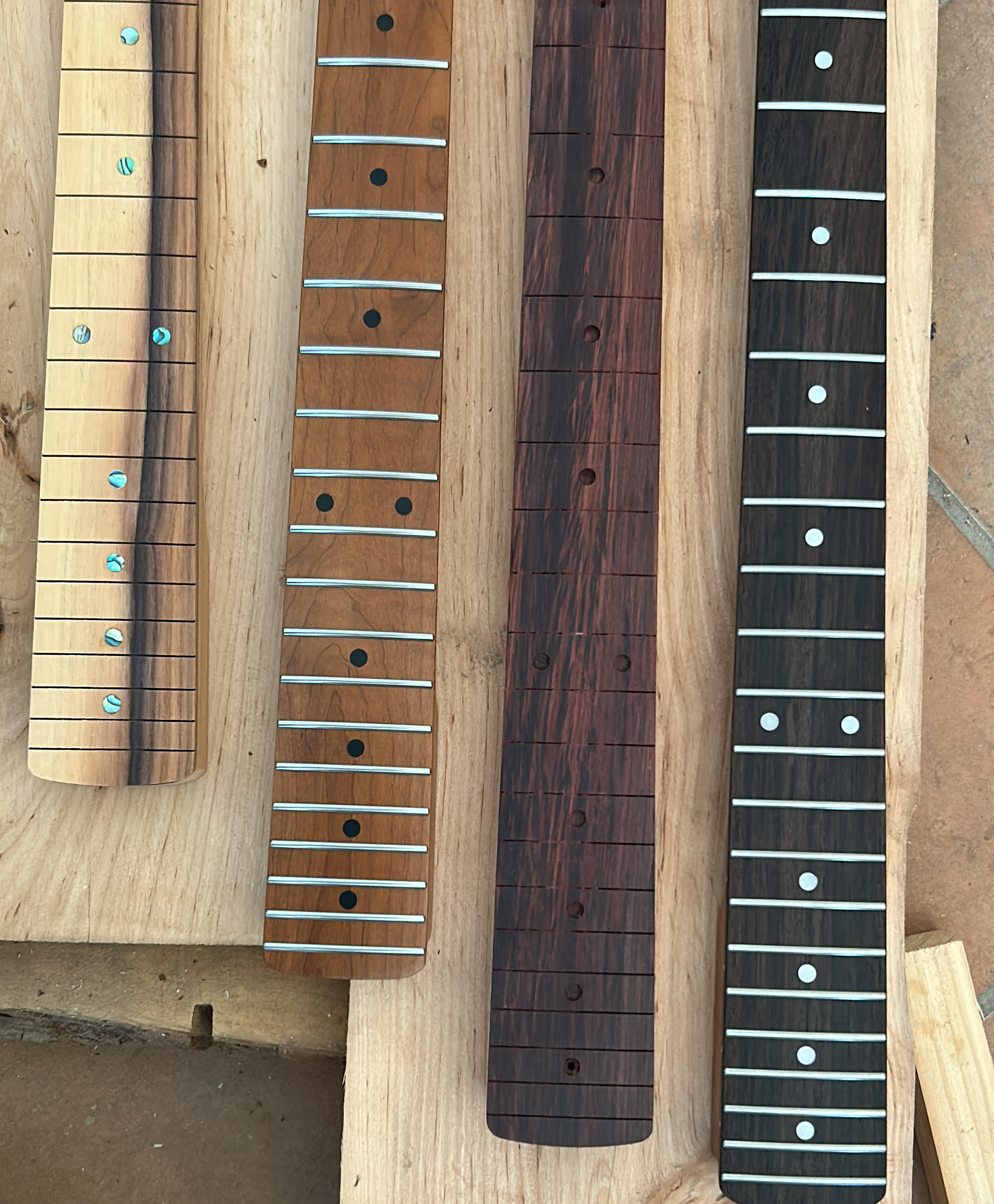 Four guitar necks or neck blanks displayed side by side on a light wood board, showing different wood finishes and inlay styles.