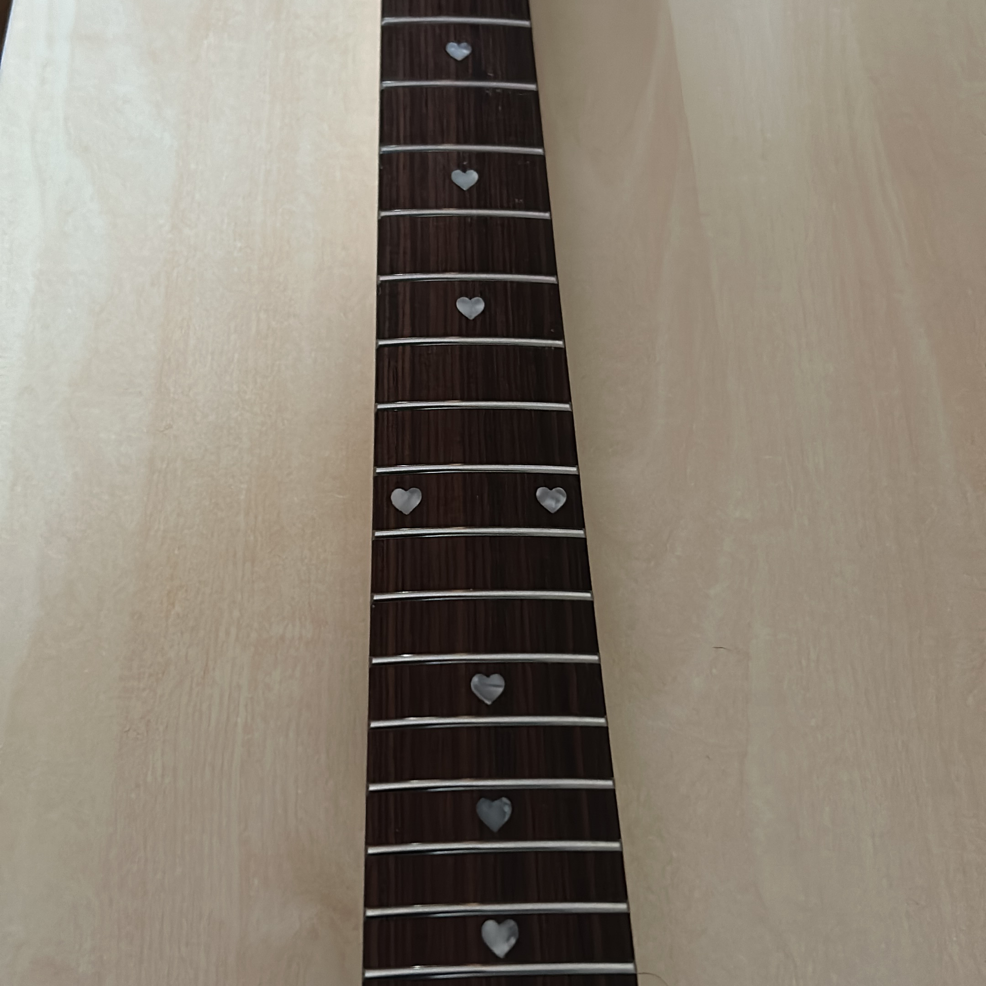 Close-up of a guitar fretboard with dark brown wood, silver metal frets, and silver heart-shaped inlays, shown against a light wood background.