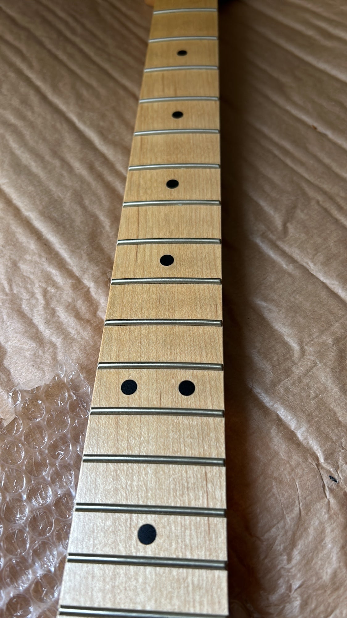 A close-up of a light natural wood guitar neck fretboard with black dot inlays, photographed diagonally on a brown textured fabric background.