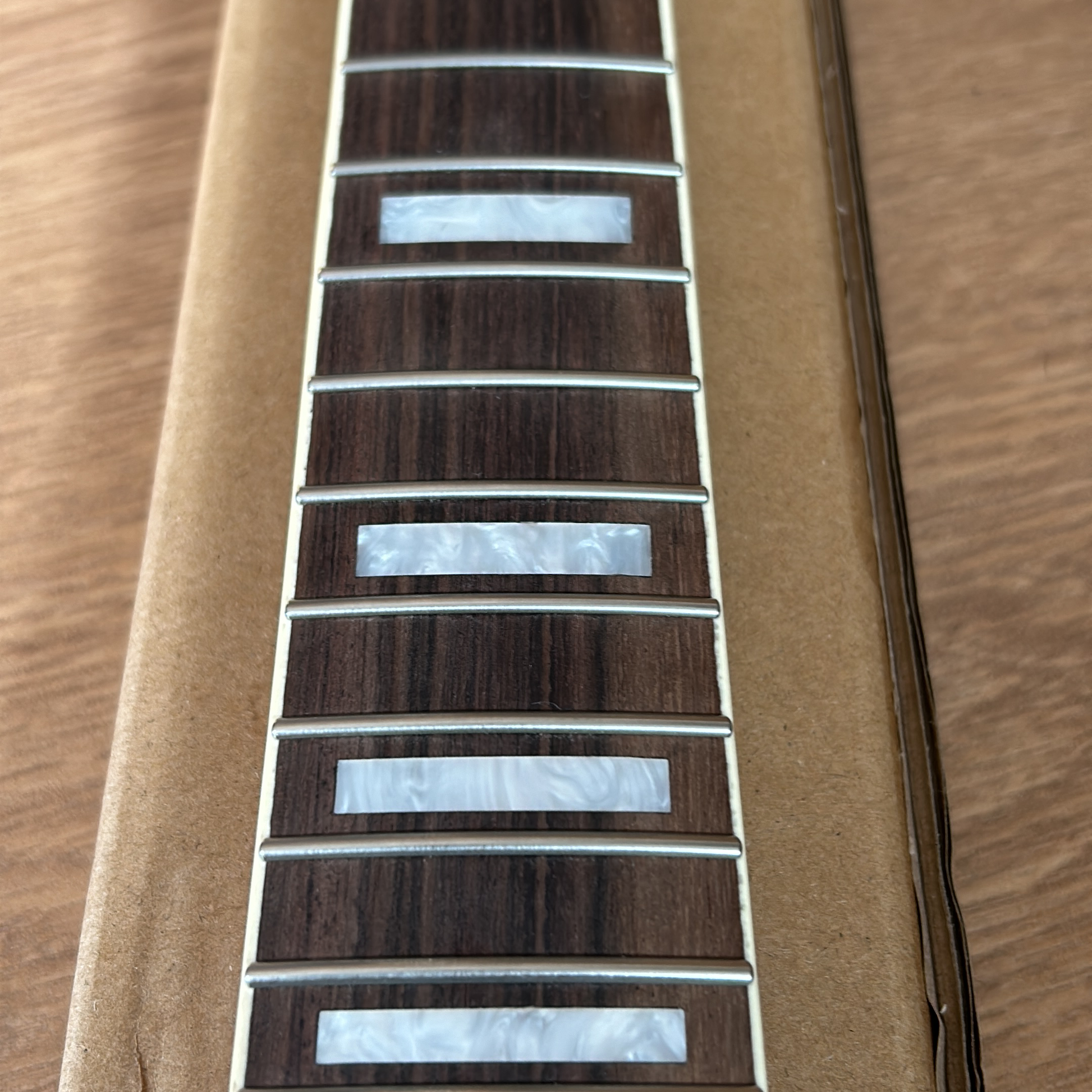 Close-up of a guitar neck fretboard with dark brown wood, silver metal fret edges, and white mother-of-pearl inlays, resting on a tan cardboard insert over a wood surface.
